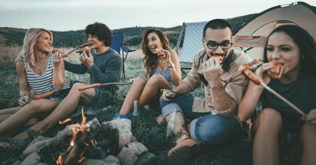 group of friends sitting around a campfire roasting sausages