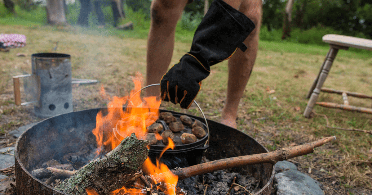 a man wearing a glove taking a pot off the campfire